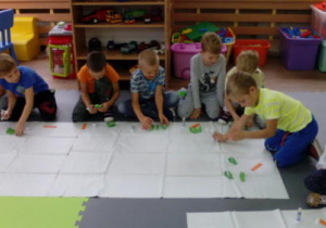 Dzieci na dywanie zgromadzone wokół ceratek, na których mają przygotowane elementy do wykonania przestrzennej marchewki.